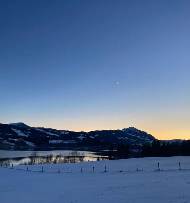 Sonnenuntergang am Rottachspeichersee im Winter, der Mond geht auf mit einer Sichel